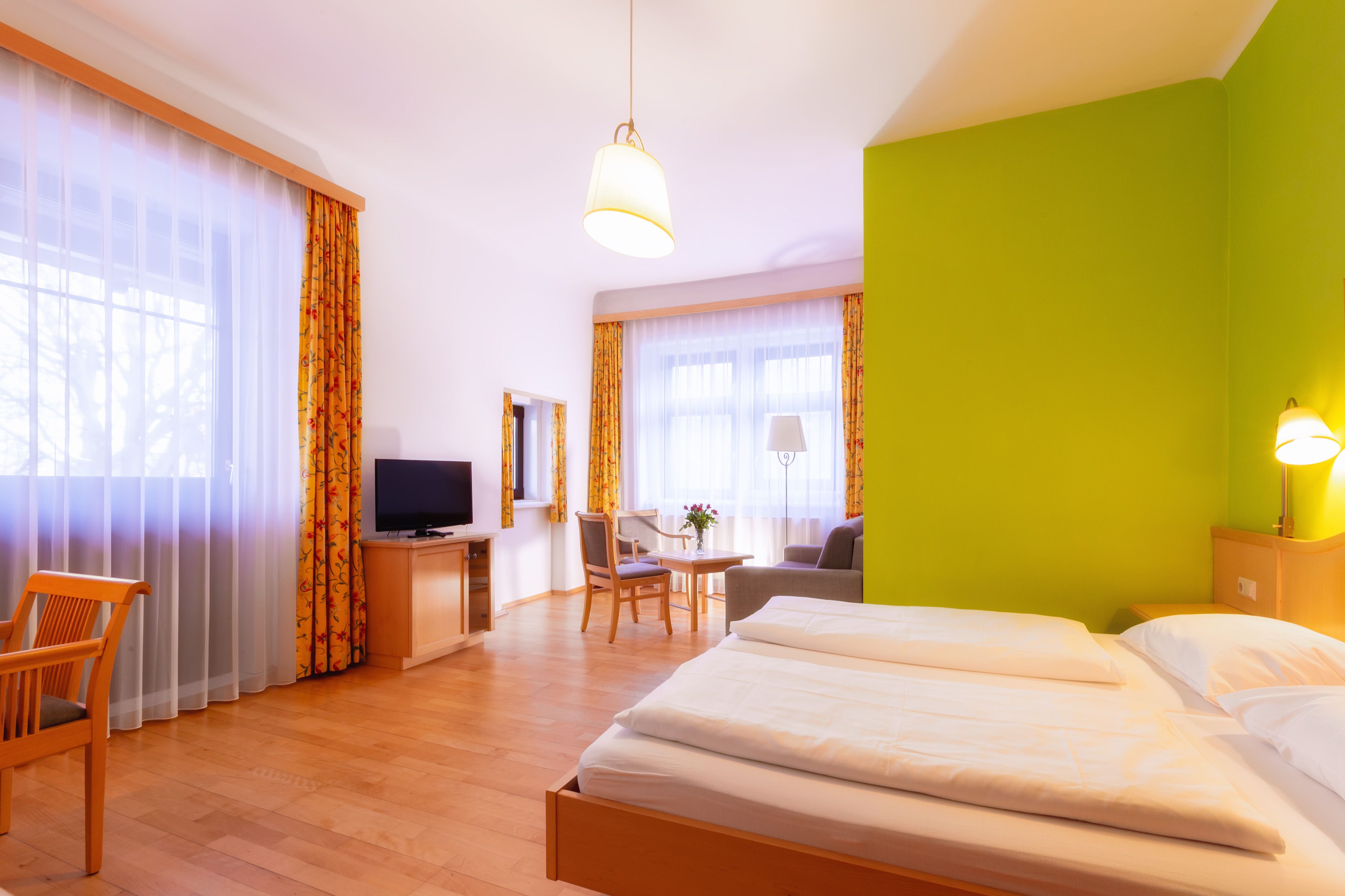 Helles Hotelzimmer mit Doppelbett, grünem Akzent an der Wand, Fernseher, Sitzbereich und großen Fenstern mit Vorhängen.