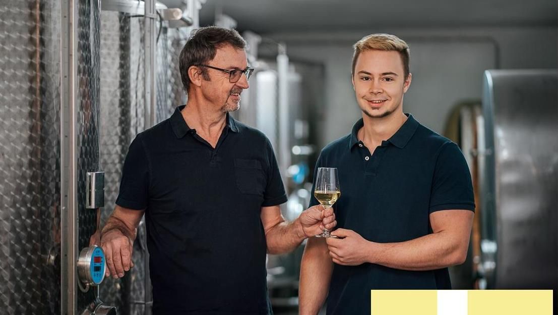 Zwei Männer in einem Weinkeller, einer hält ein Glas Weißwein, der andere bedient einen Tank.