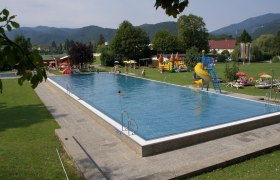 Ein Freibad mit Schwimmbecken, Rutsche und Liegewiese, umgeben von Bäumen und Bergen.