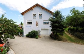 Ein zweist&ouml;ckiges Haus mit blauer Fassade und Holzdach, umgeben von B&auml;umen und einem gepflasterten Weg.