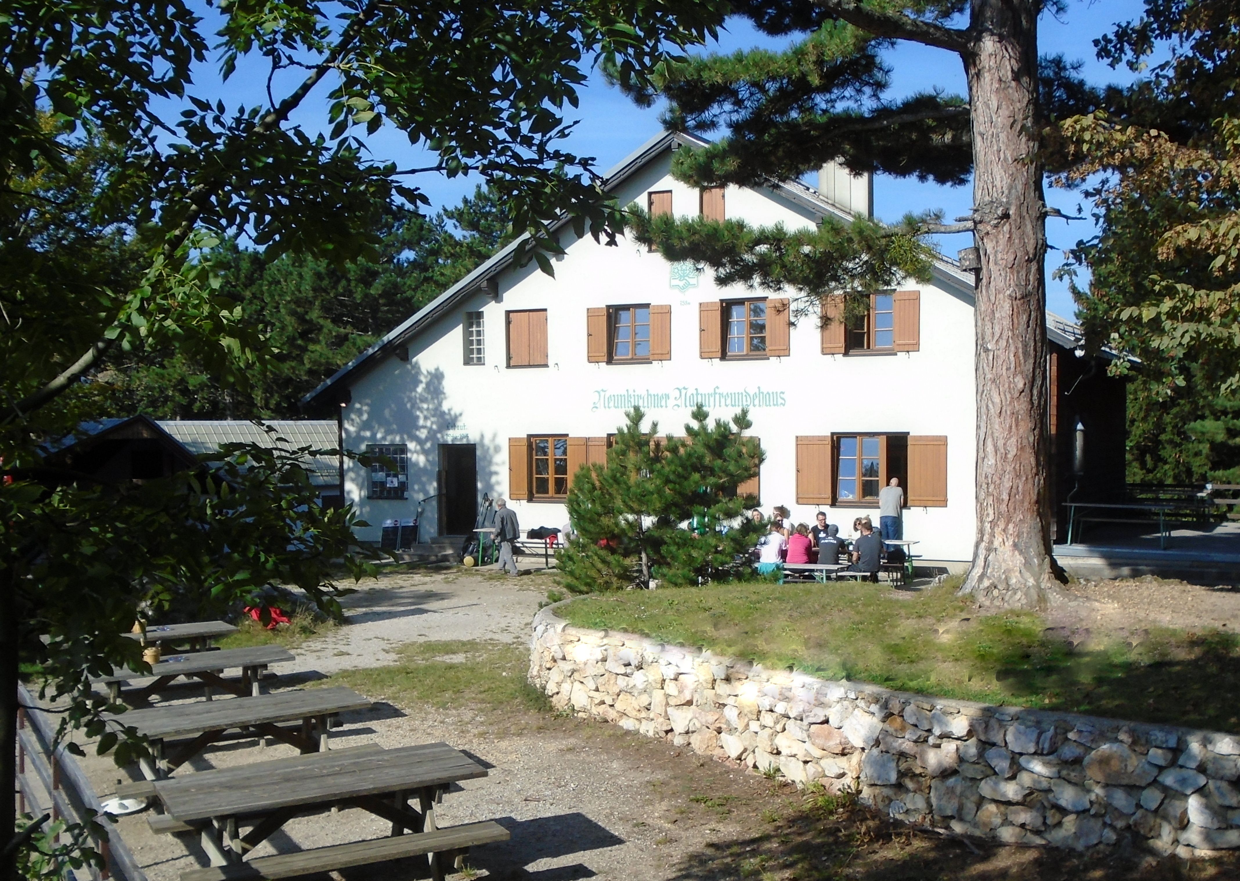 Ein Gebäude mit der Aufschrift 'Neunkirchner Naturfreundehaus', umgeben von Bäumen und Sitzbänken im Freien.