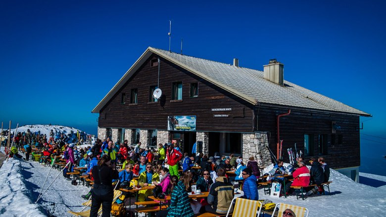 Menschen sitzen vor einem Berghaus im Schnee, blauer Himmel.