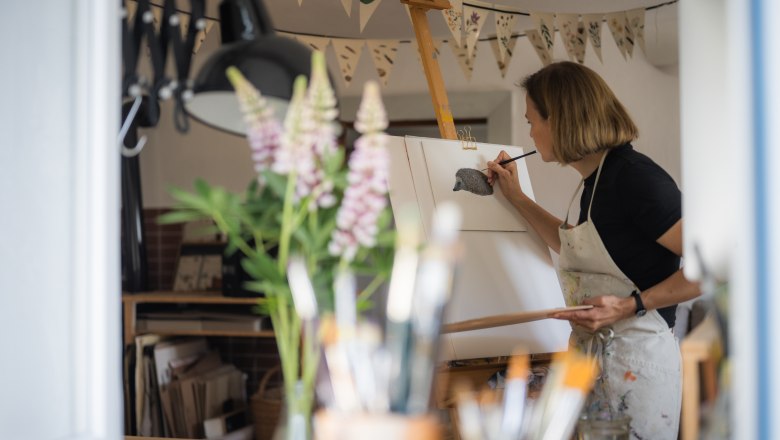 Eine Frau malt in einem Atelier an einer Staffelei ein Bild von einem Igel. Im Vordergrund sind Pinsel und Blumen zu sehen.