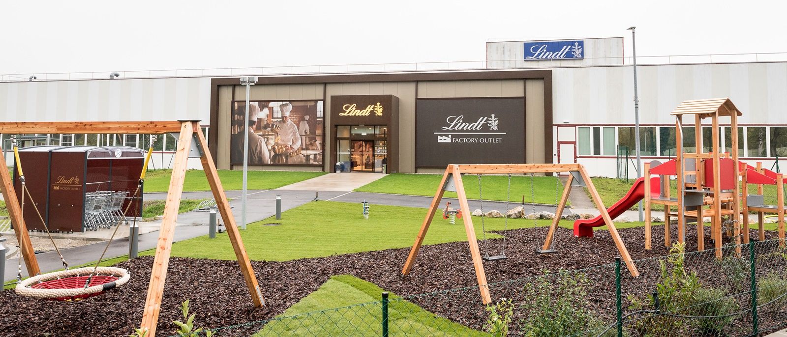 Spielplatz vor einem Lindt Factory Outlet mit Schaukeln und Rutsche.