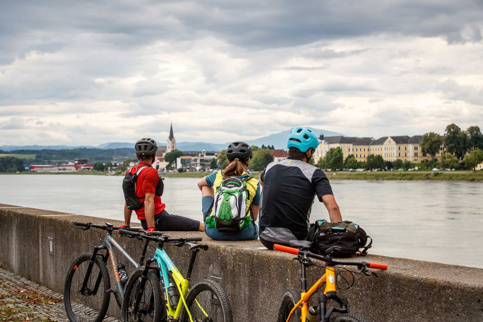 Drei Radfahrer sitzen auf einer Mauer am Flussufer und blicken auf eine Stadt mit Kirche und Gebäuden im Hintergrund.