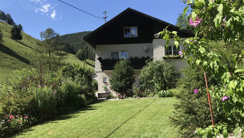 Außenansicht, © Familie Krenn Ein zweistöckiges Haus mit Balkon, umgeben von einem gepflegten Garten und grünen Hügeln im Hintergrund.