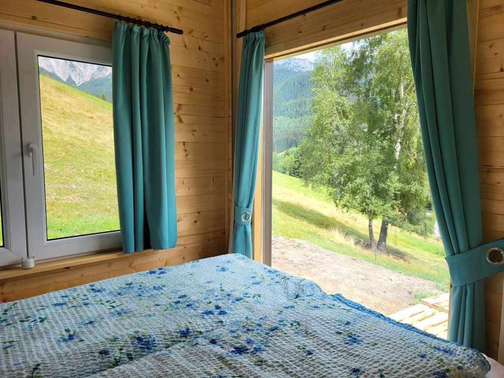 Innenansicht eines Zimmers mit Bett und großen Fenstern, die einen Blick auf eine grüne Landschaft bieten.