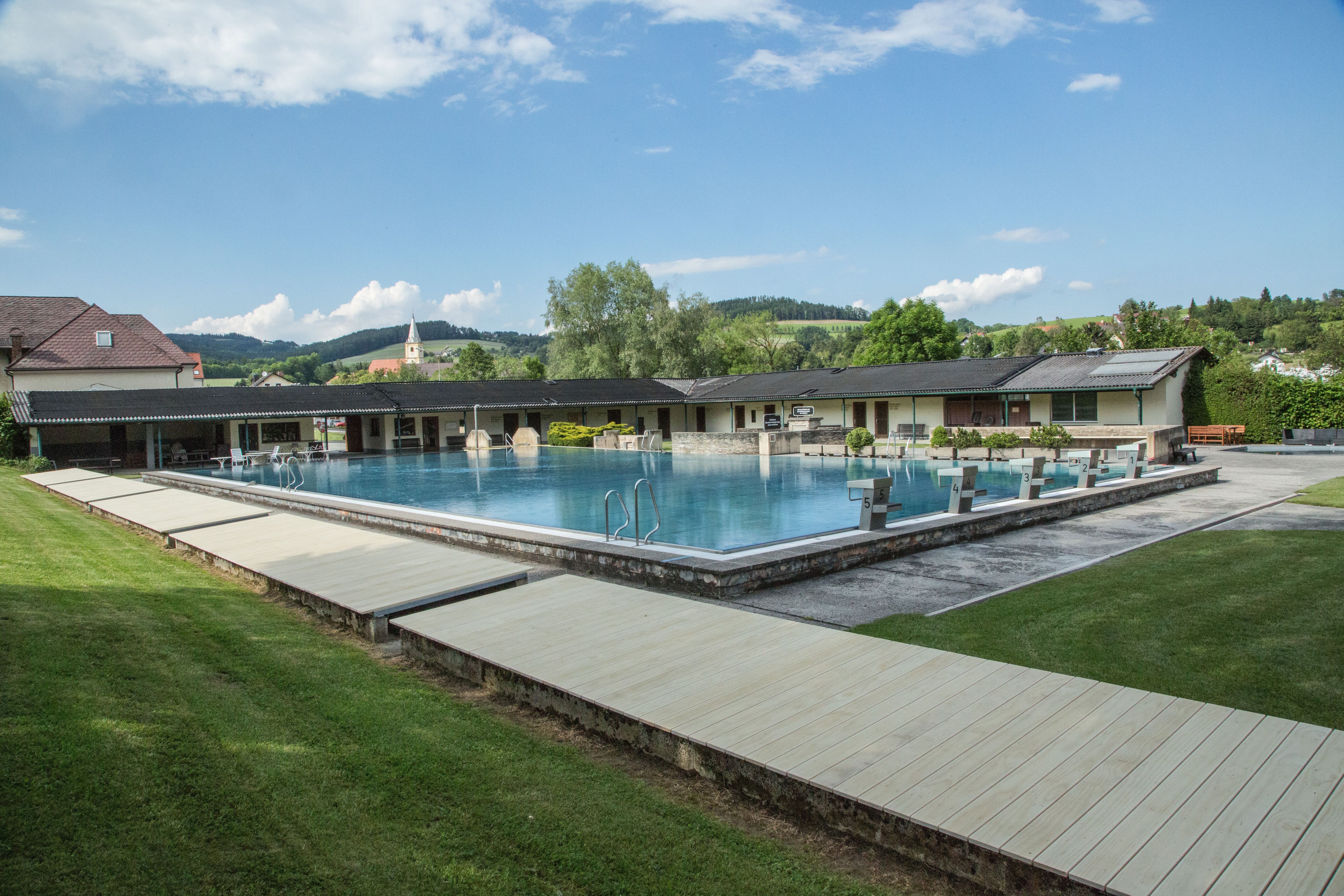 Freibad Krumbach mit Schwimmbecken und Sprungblöcken, umgeben von grüner Landschaft und Gebäuden.