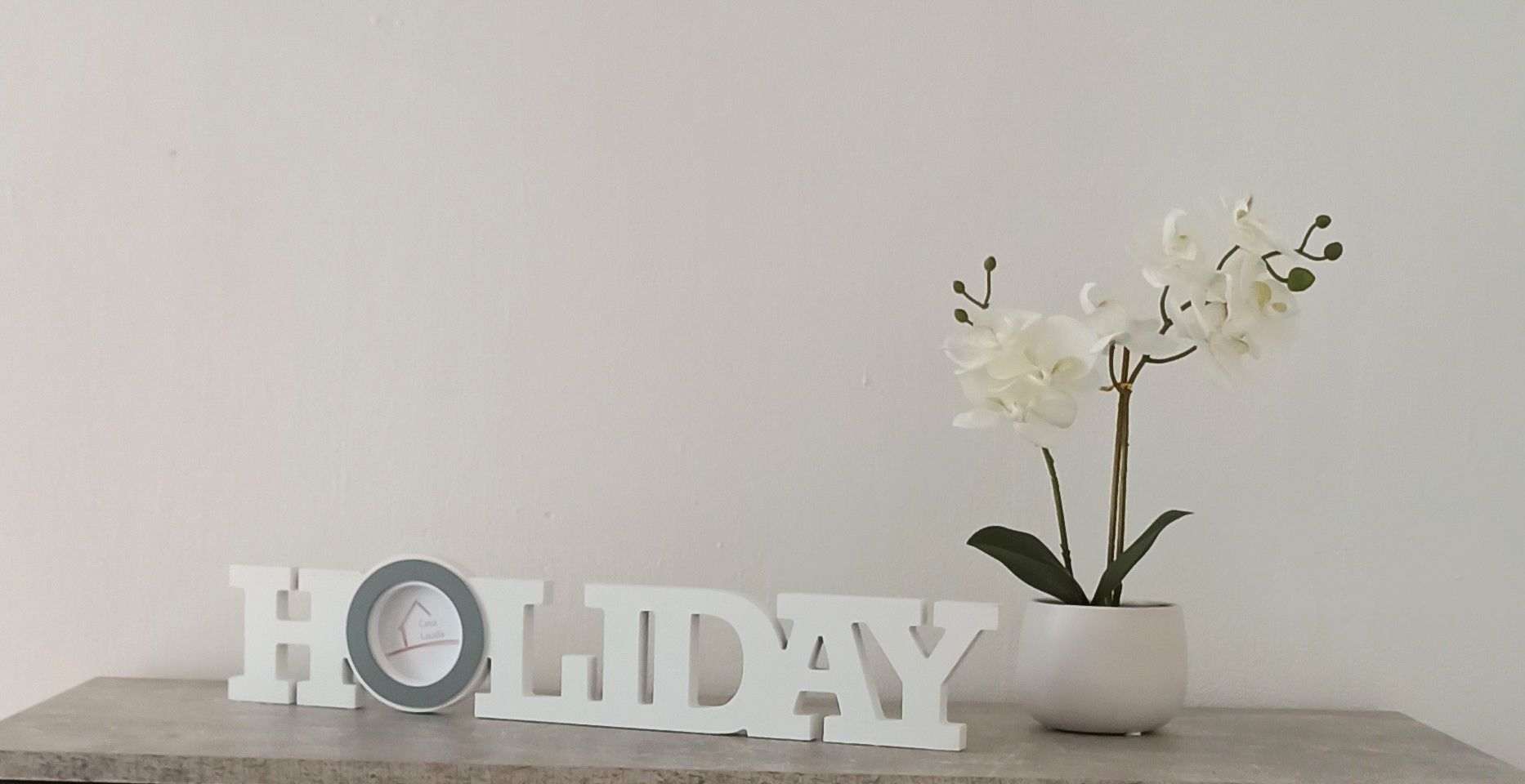 Dekoration mit Schriftzug 'HOLIDAY' und weißer Orchidee in einem Topf auf einem Tisch.