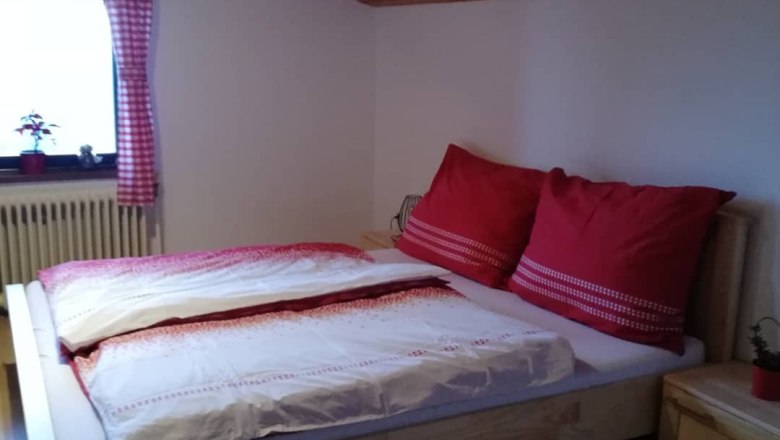 Gem&uuml;tliches Schlafzimmer mit Holzbett, roten Kissen und karierten Vorh&auml;ngen unter einer Holzdecke.