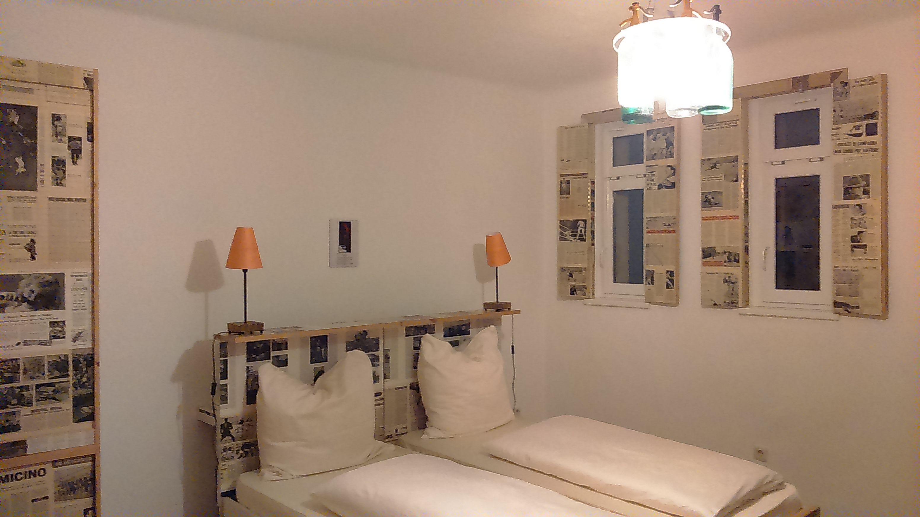 Hotelzimmer mit Doppelbett, Zeitungsdekoration an Wänden und Fenstern, zwei orangefarbene Lampen.
