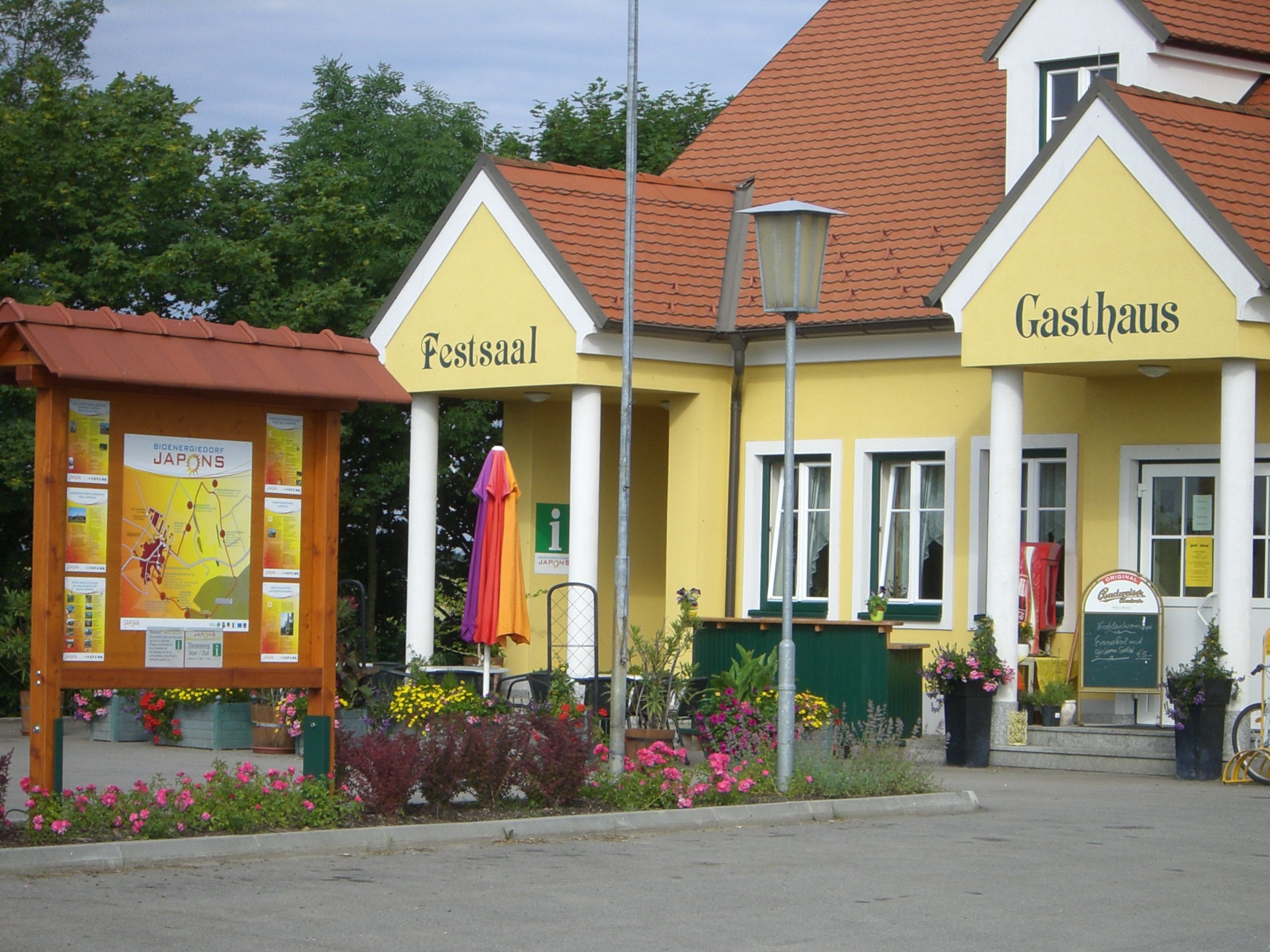 Gelbes Gebäude mit der Aufschrift 'Festsaal' und 'Gasthaus', umgeben von Blumen und einem Informationsschild.