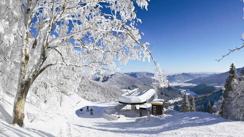 Winterlandschaft mit Sesselliftstation, verschneiten Bäumen und Bergen im Hintergrund.