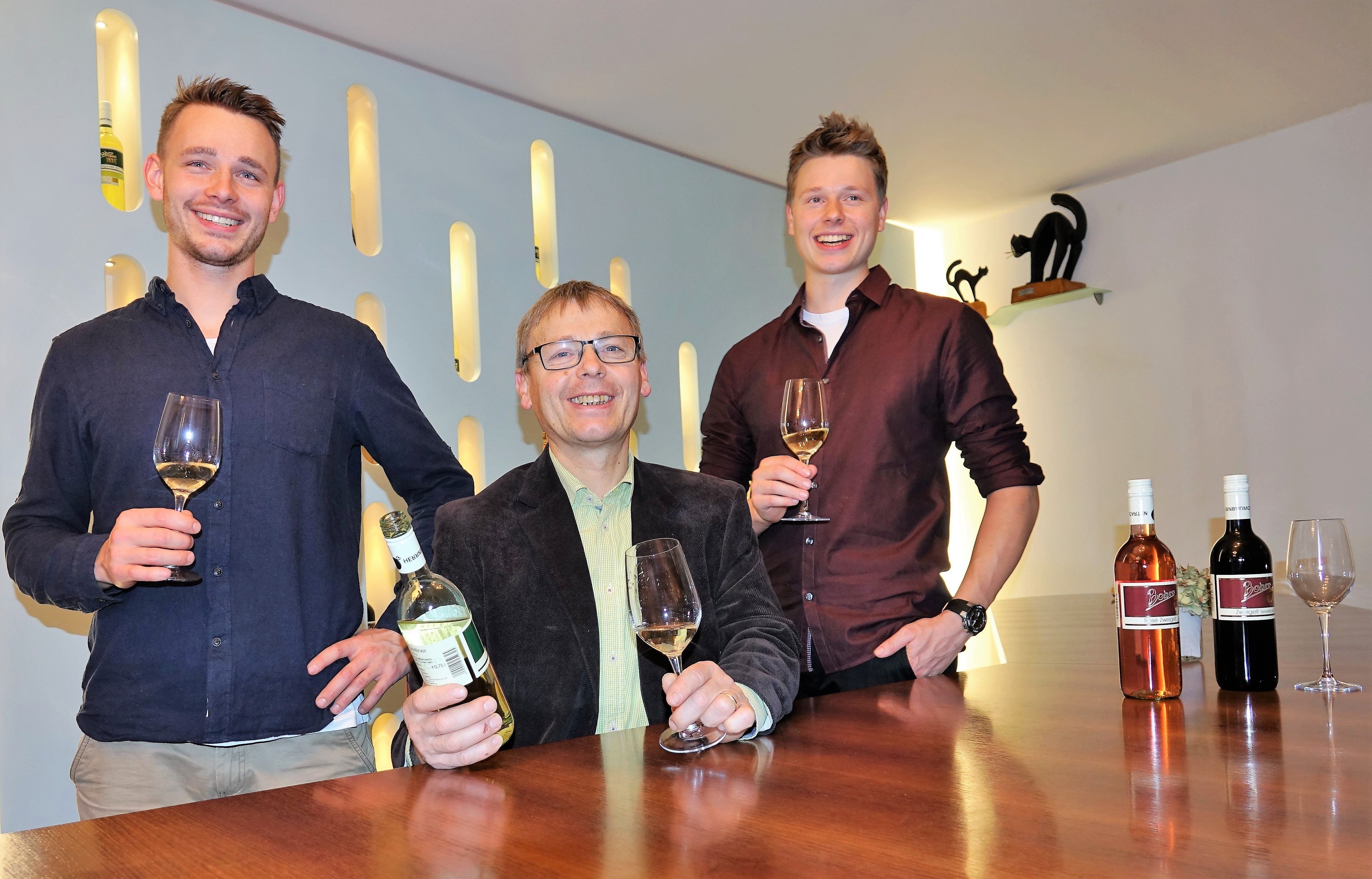 Drei Männer stehen an einem Tisch mit Weingläsern und Weinflaschen.