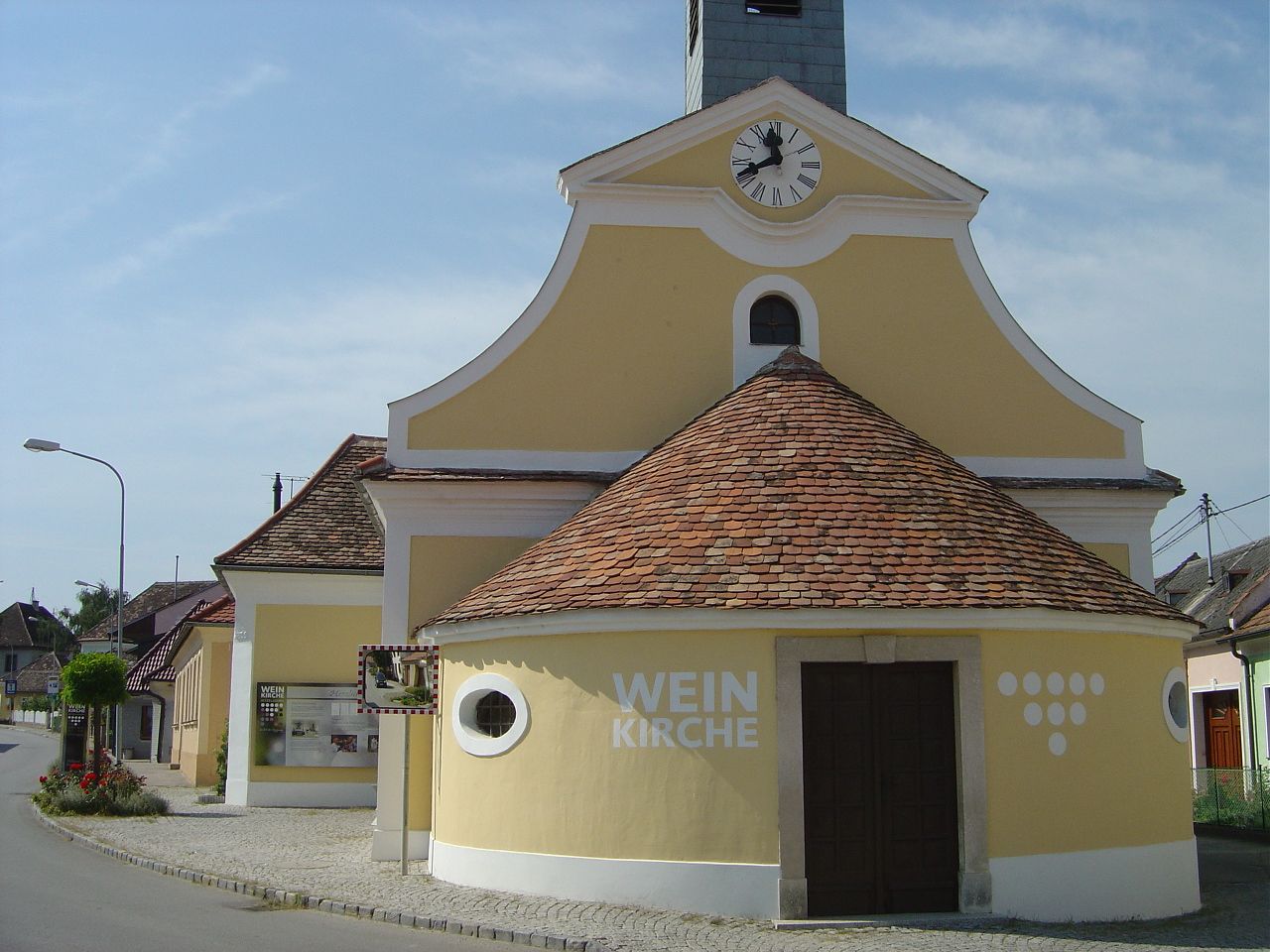 Gelbe Kirche mit Uhrturm und der Aufschrift 'Weinkirche' in einem Dorf.