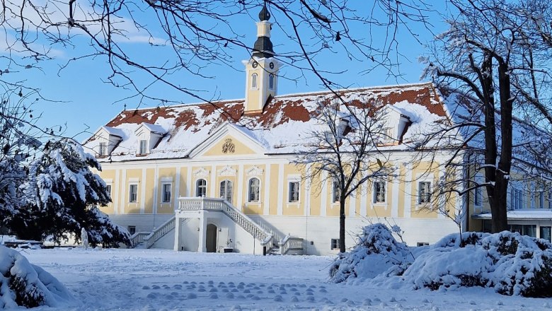 Schloss Haindorf im Winter mit Schnee bedeckt, blauer Himmel und kahle B&auml;ume im Vordergrund.