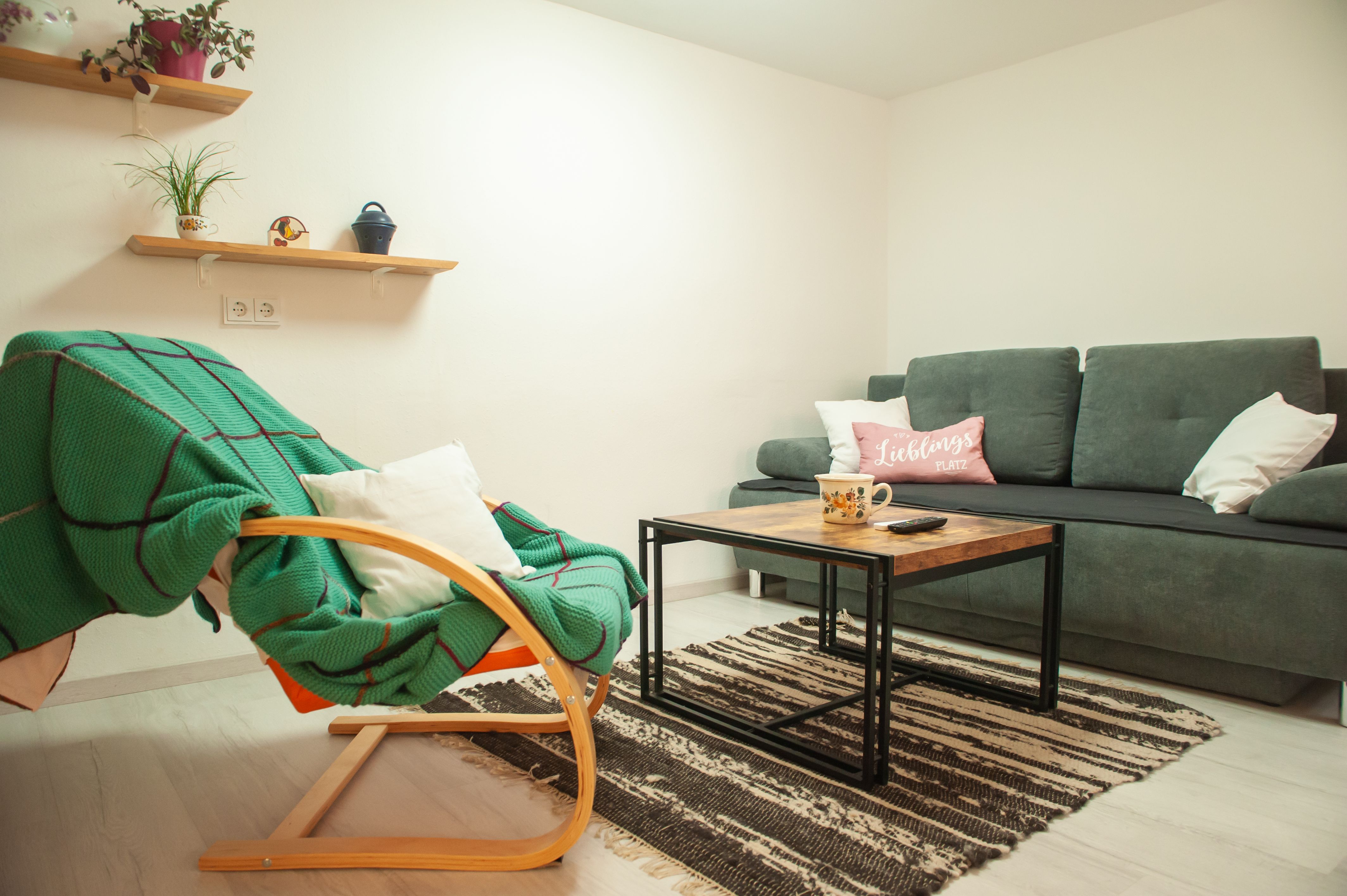 Gemütliches Wohnzimmer mit grauem Sofa, Holztisch, grünem Schaukelstuhl und Regalen mit Pflanzen.