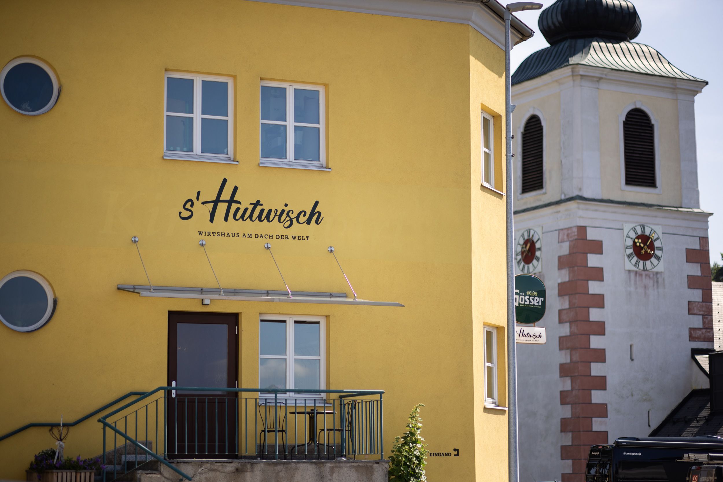 Gelbes Gebäude mit der Aufschrift 's'Hutwisch' neben einem Kirchturm mit Uhr.