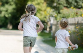 Zwei Kinder laufen auf einem Kiesweg in einem Park, umgeben von B&auml;umen und Blumen.