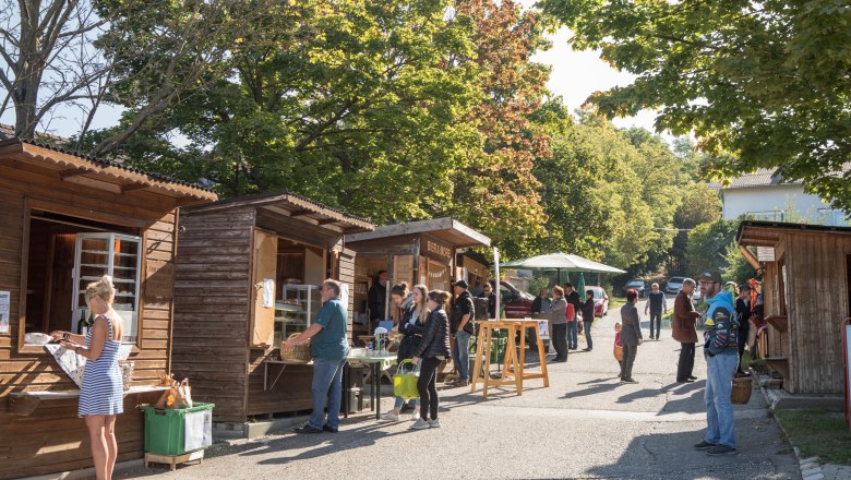 Menschen stehen an Holzh&uuml;tten auf einem Markt im Freien bei sonnigem Wetter.
