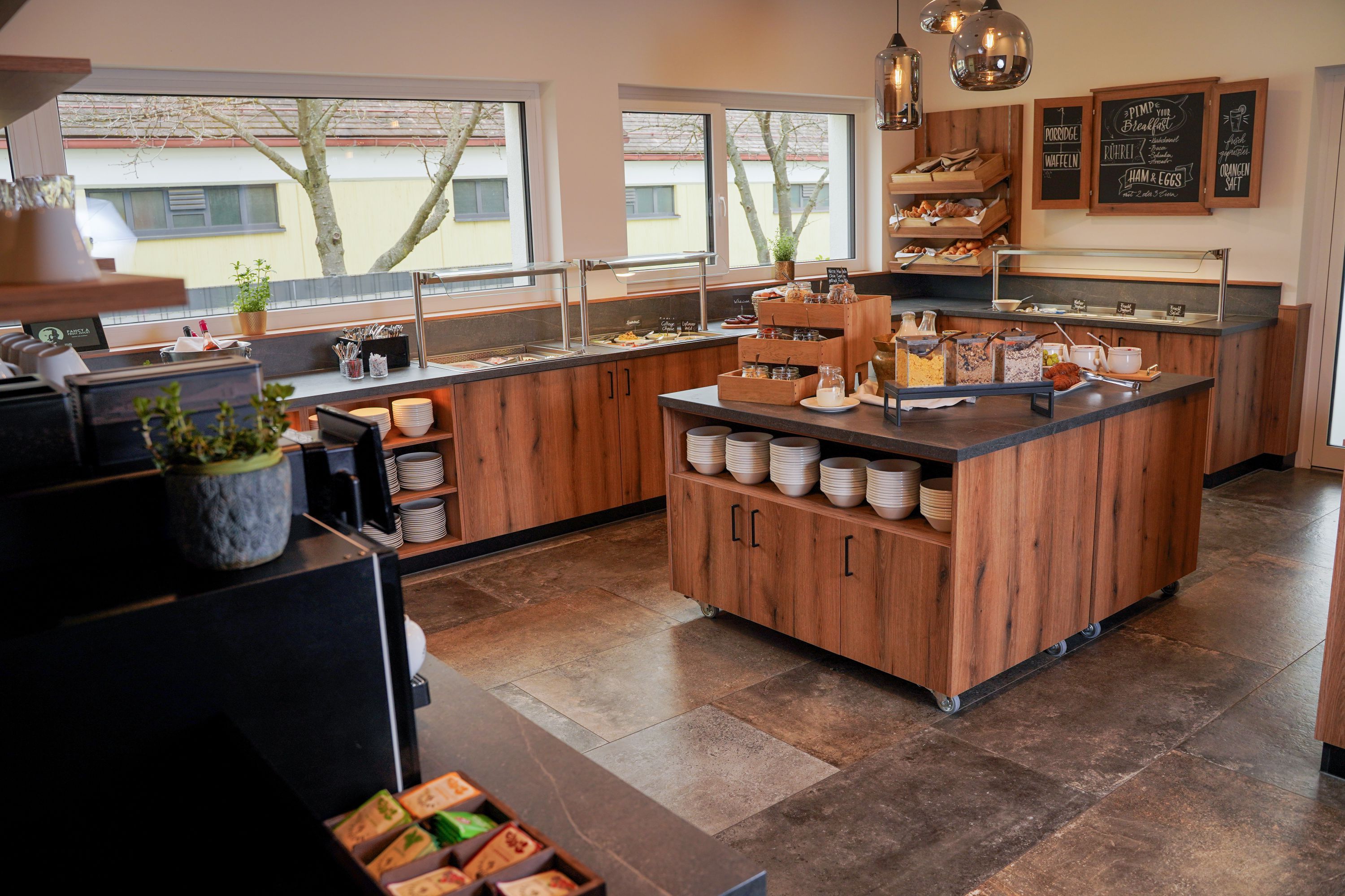 Frühstücksraum mit Buffet, Holztheke, Schalen, Müsli, Gebäck und großen Fenstern. Der Titel Egino passt zum gezeigten Raum nicht eindeutig.