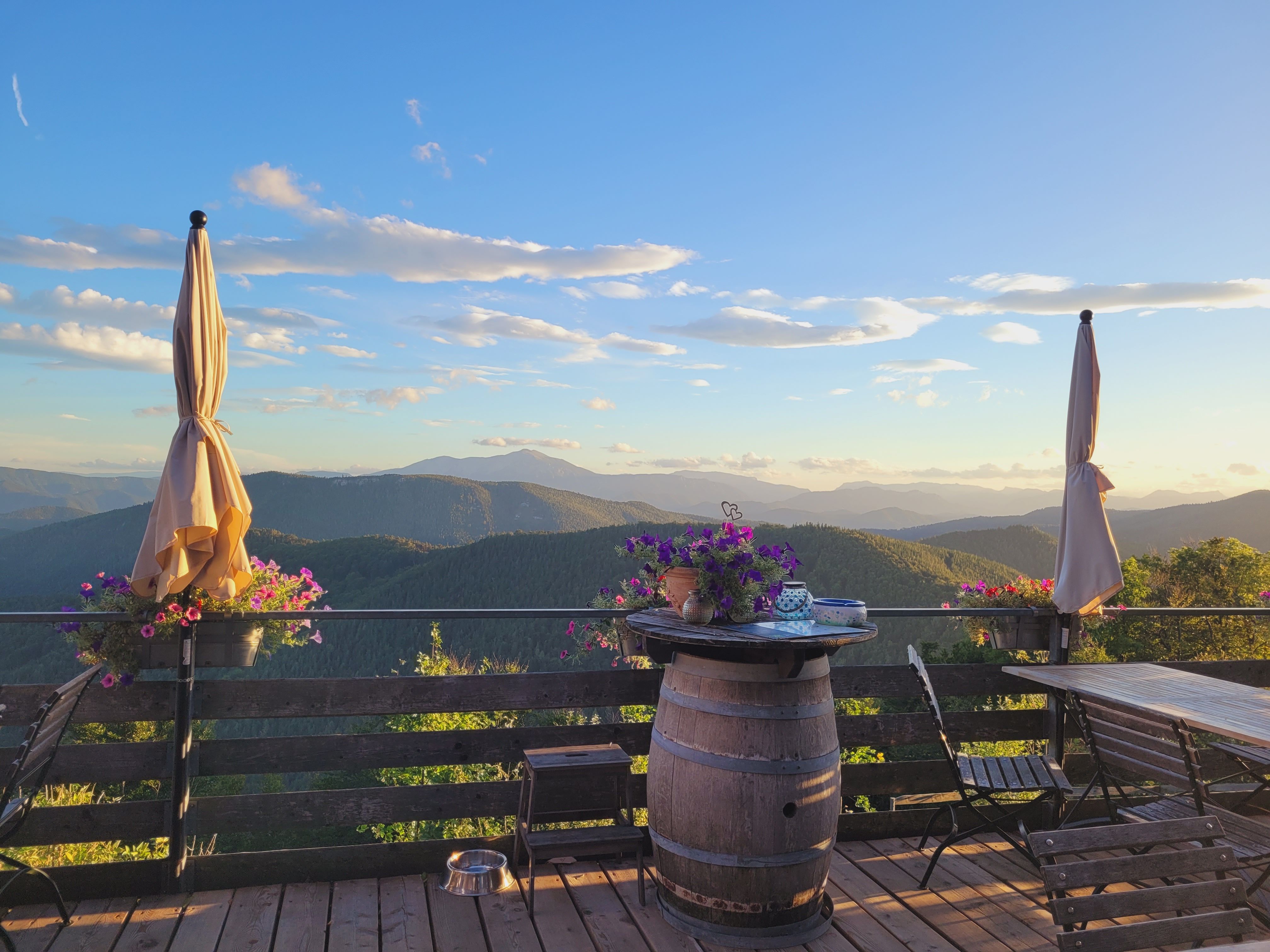 Terrasse der Enzianhütte mit Blick auf Berglandschaft bei Sonnenuntergang, zwei geschlossene Sonnenschirme, Blumen und Holztisch.