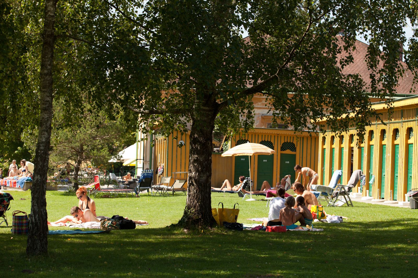 Menschen entspannen auf einer Wiese vor einem historischen Badehaus.
