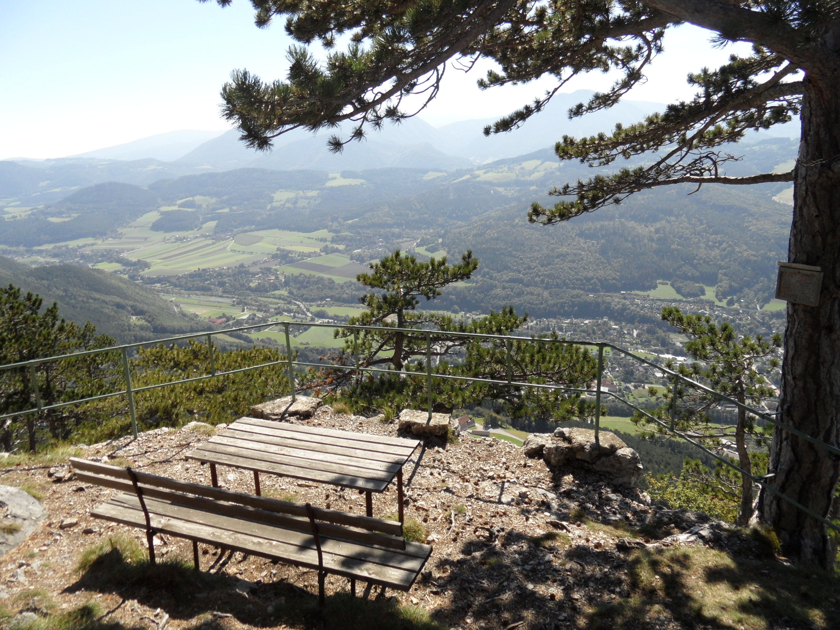 Aussichtspunkt mit Bank und Tisch, umgeben von Bäumen, mit Blick auf ein Tal und Berge im Hintergrund.