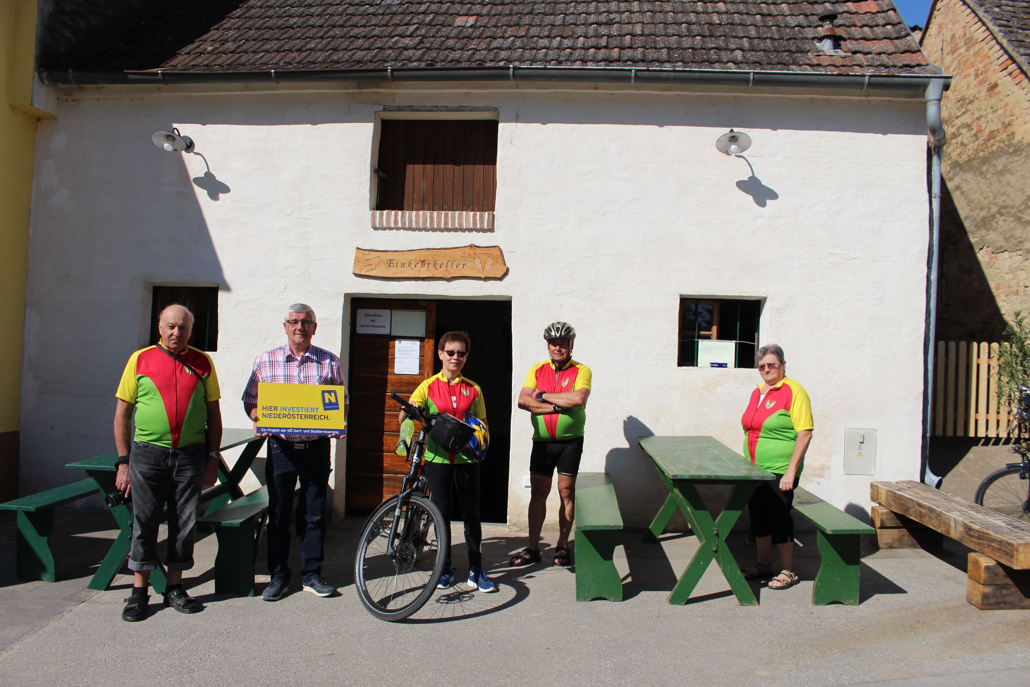 Eine Gruppe von fünf Personen steht vor dem Einkehrkeller Ottenthal. Zwei Personen tragen Radtrikots, eine hält ein Fahrrad. Ein Schild weist auf Investitionen in Niederösterreich hin.