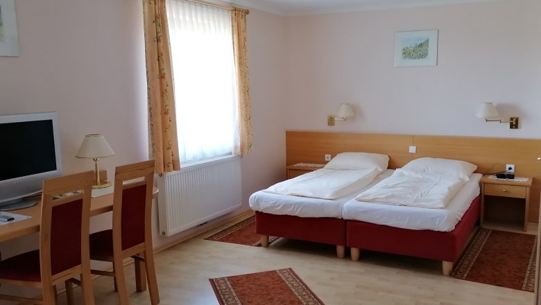 Ein Doppelzimmer mit zwei Einzelbetten, einem Schreibtisch mit St&uuml;hlen, einem Fernseher und einem Fenster mit Vorh&auml;ngen.