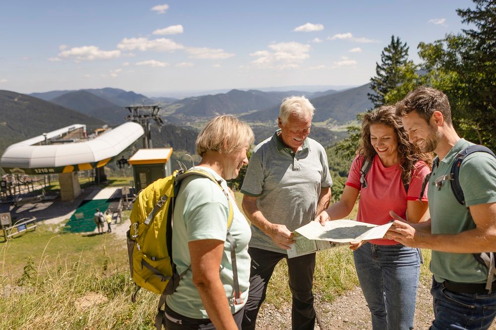 Vier Personen betrachten eine Karte vor einer Bergbahnstation in einer bergigen Landschaft.