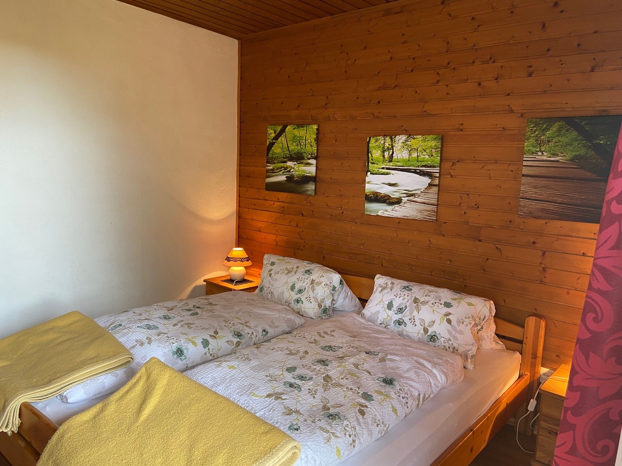 Gemütliches Doppelzimmer mit Holzwänden, einem Doppelbett mit Blumenbettwäsche und gelben Decken, sowie einer Nachttischlampe.