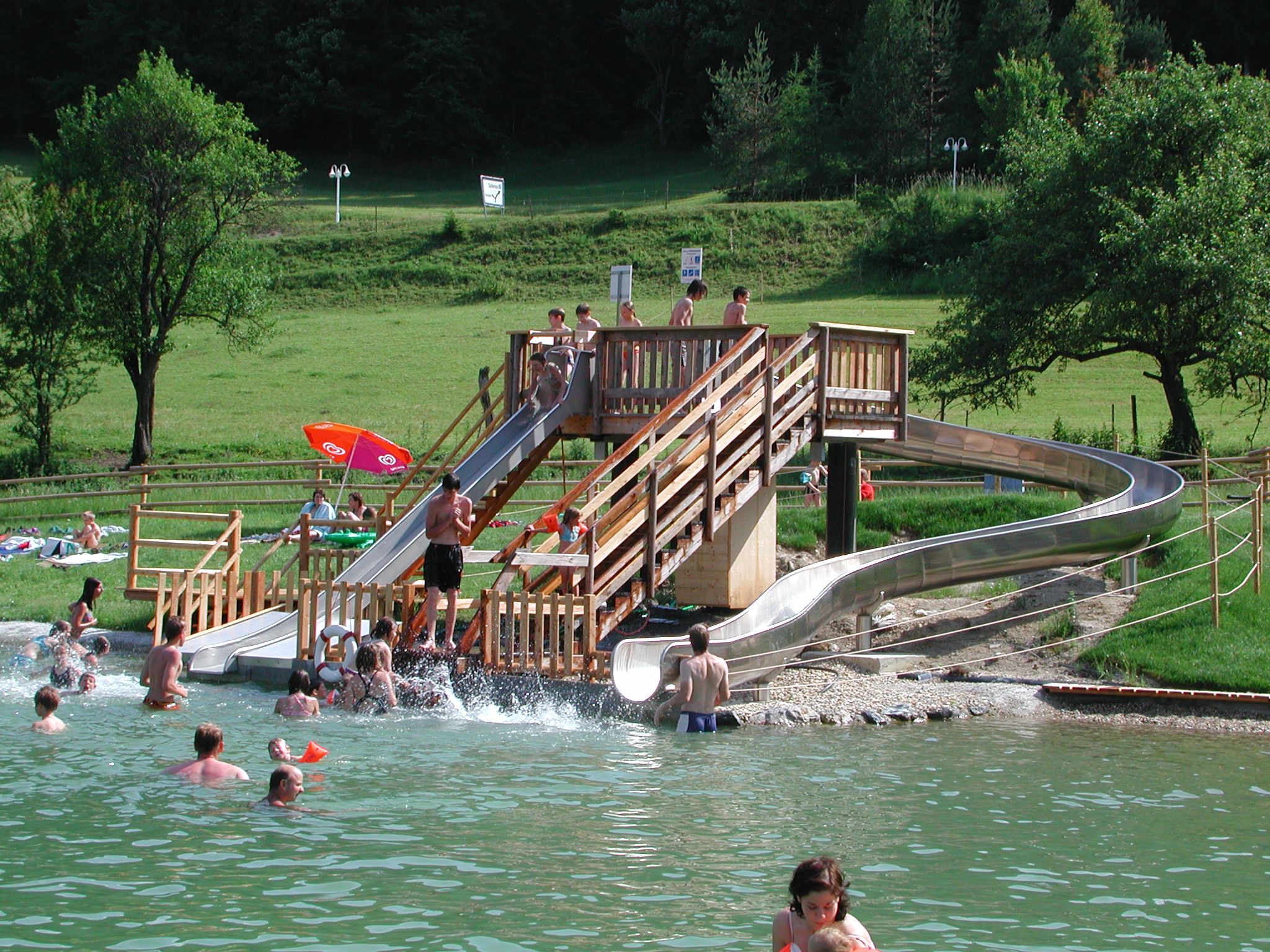 Menschen genießen eine Wasserrutsche in einem Naturbad mit grüner Umgebung.