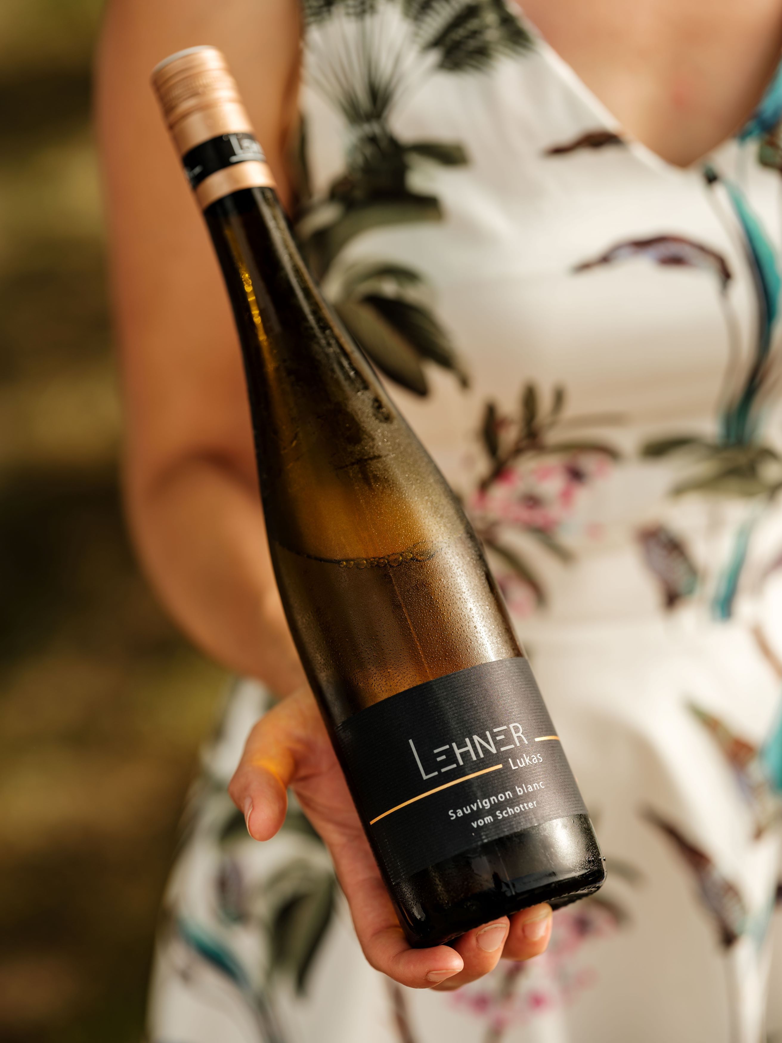 Eine Person hält eine Weinflasche mit dem Etikett 'Lehner Lukas Sauvignon blanc vom Schotter'.