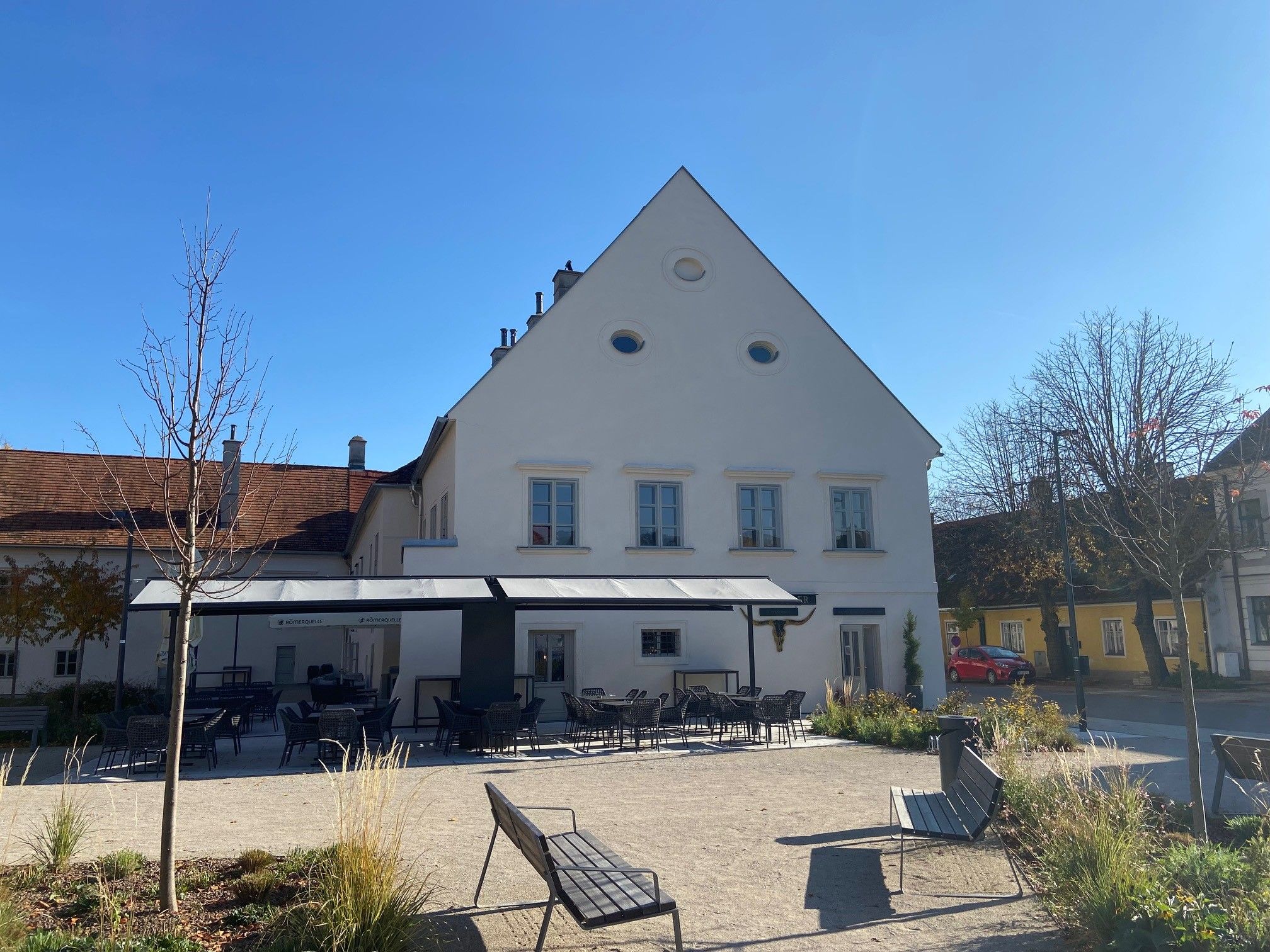Ein Gastgarten mit leeren Tischen und Stühlen vor einem weißen Gebäude mit spitzem Dach an einem sonnigen Tag.