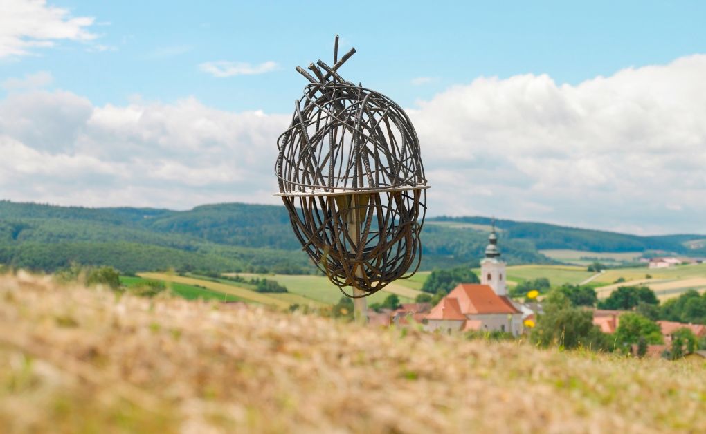 Kunstinstallation aus Metall in einer ländlichen Landschaft mit Kirche im Hintergrund.
