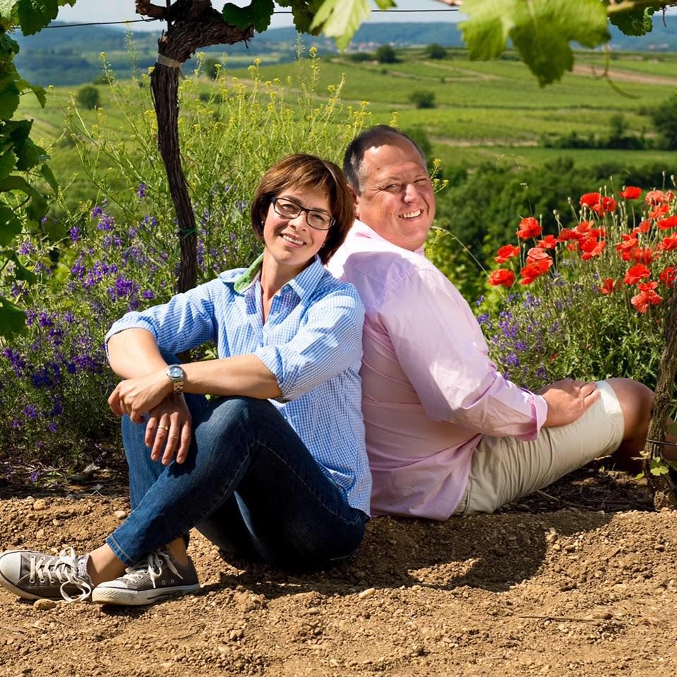 Zwei Personen sitzen Rücken an Rücken in einem Weinberg mit Blumen im Hintergrund.