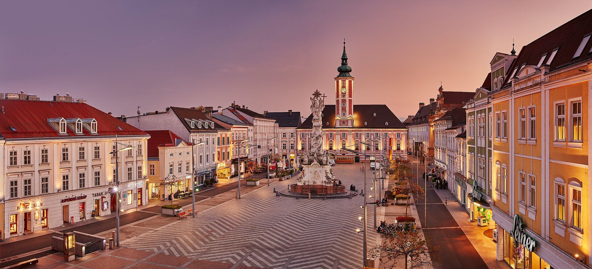 Die malerische Kulisse der Stadt erstrahlt im warmen Licht der Abenddämmerung. Historische Gebäude umrahmen den zentralen Platz, während die sanften Farben des Himmels eine einladende Atmosphäre schaffen. Hier trifft Tradition auf modernes Leben und lädt zum Verweilen ein.