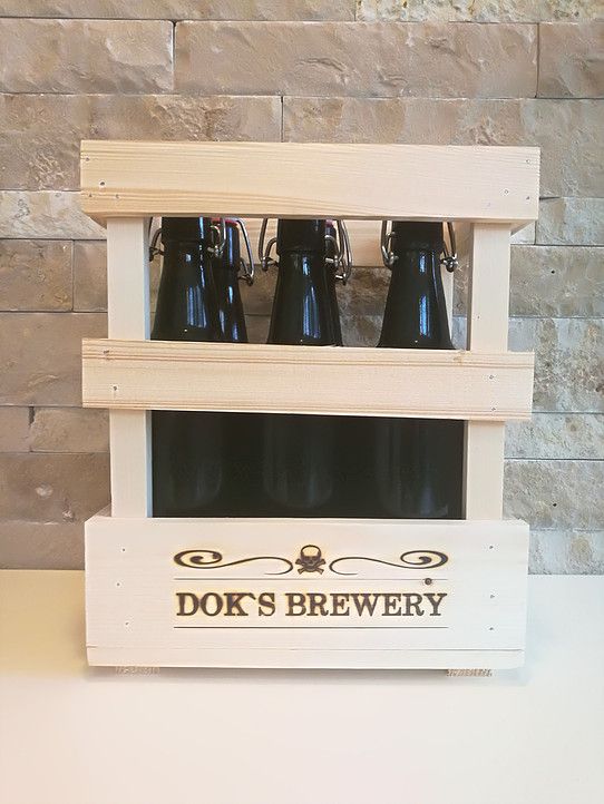 Holzkiste mit drei dunklen Bierflaschen von Dok's Brewery vor einer Steinwand.