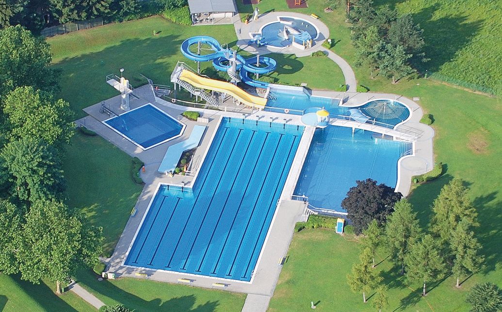 Luftaufnahme eines Freibads mit mehreren Becken und Wasserrutschen, umgeben von grünen Bäumen und Rasenflächen.