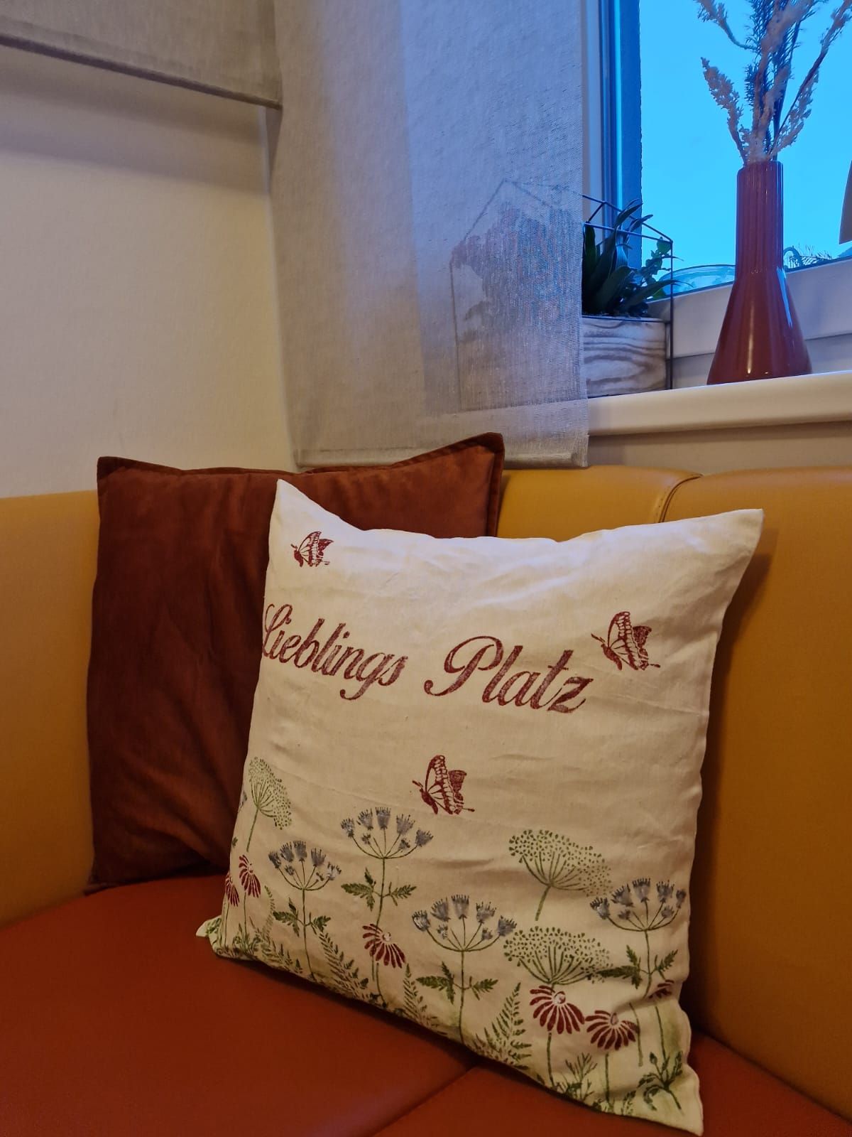 Ein gemütliches Sofa mit einem dekorativen Kissen, auf dem 'Lieblings Platz' steht, neben einem Fenster mit Pflanzen und einer Vase.