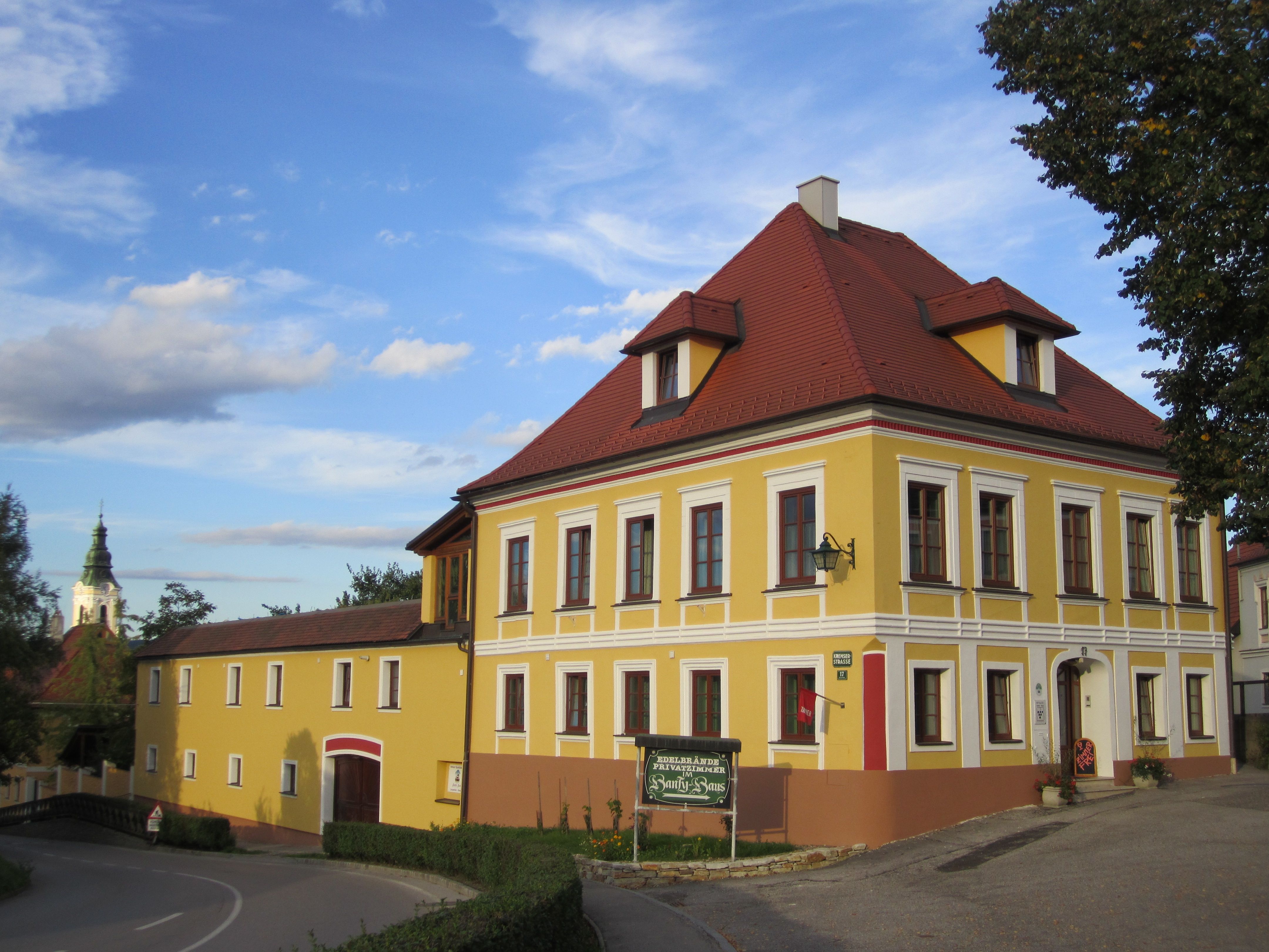 Gelbes zweistöckiges Gebäude mit rotem Dach und weißen Fensterrahmen, daneben ein kleineres Gebäude. Im Hintergrund eine Kirchturmspitze.