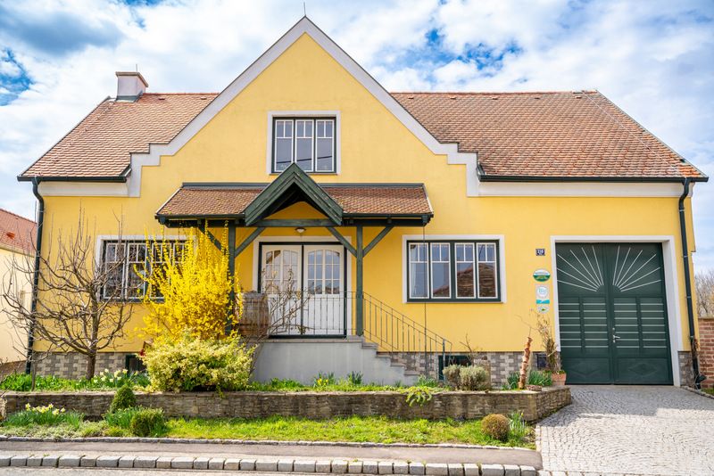 Gelbes Haus mit rotem Dach und grünem Tor, umgeben von Garten und blühenden Sträuchern.