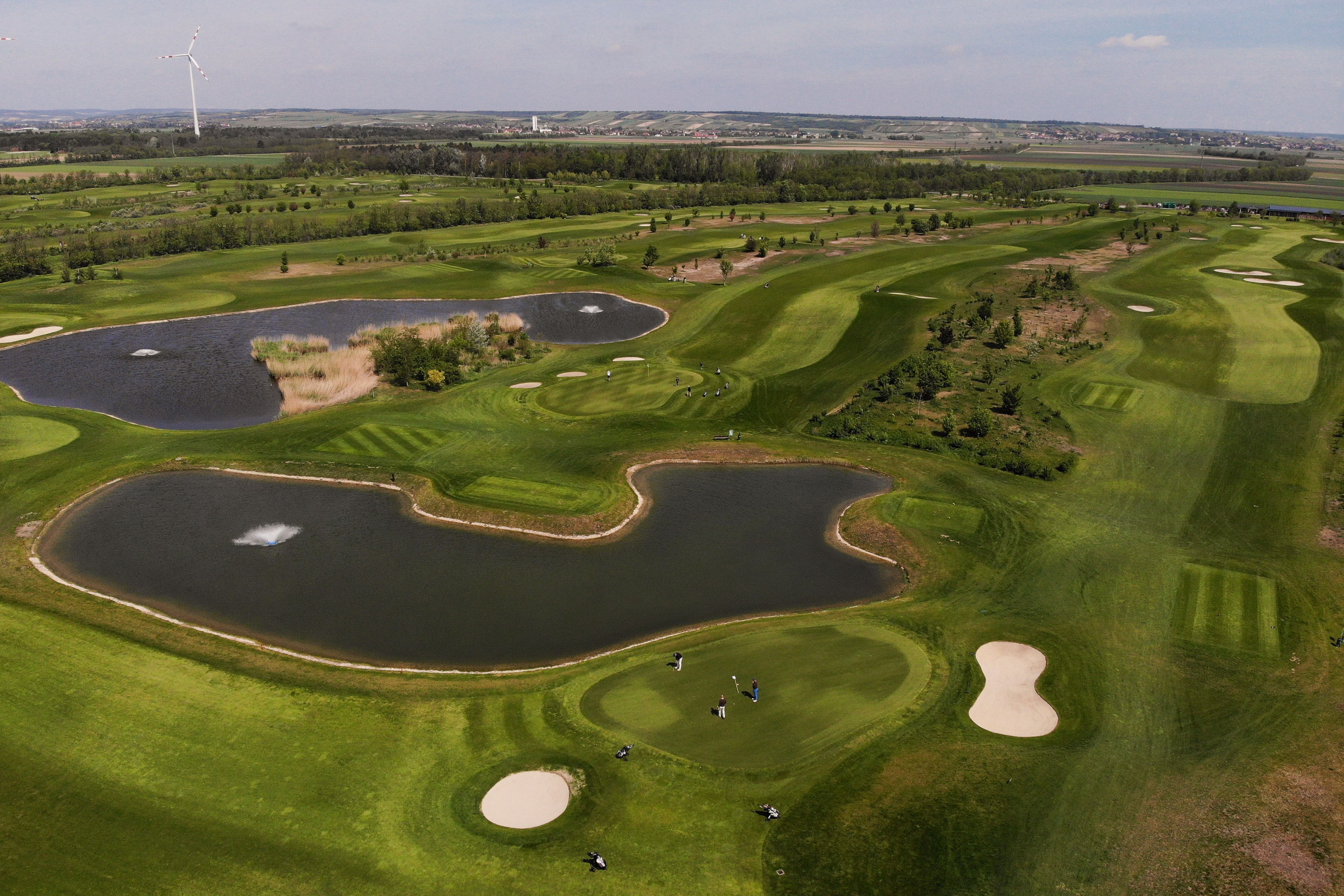 Luftaufnahme eines Golfplatzes mit Teichen und Windrad im Hintergrund.