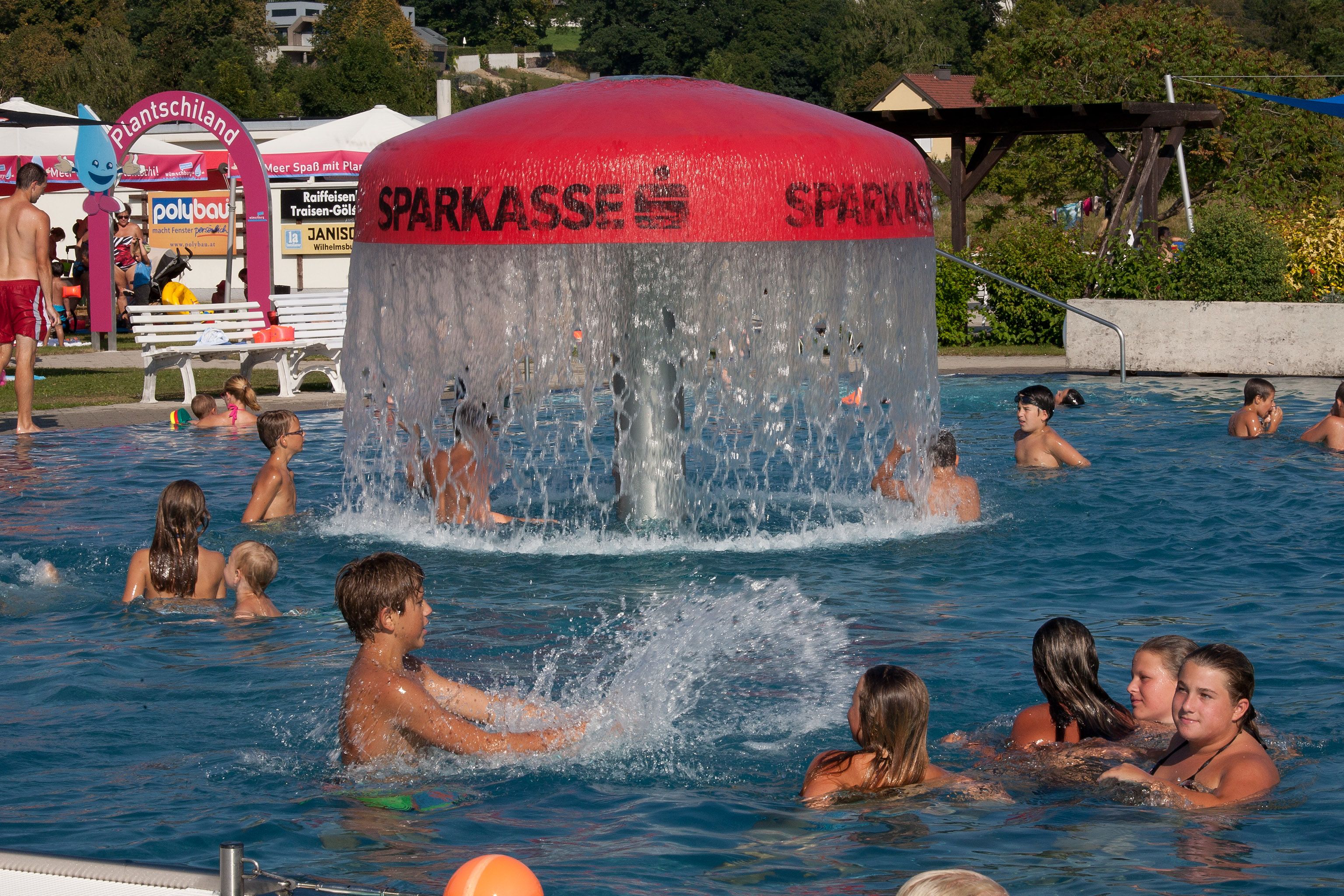 Menschen schwimmen in einem Freibad mit einem großen Wassersprudler in der Mitte.