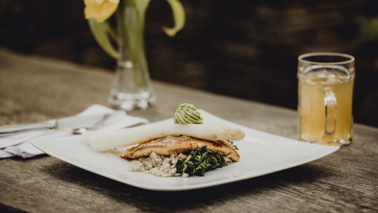 Gourmetgericht mit Spargel, Fisch und Gem&uuml;se auf Holztisch, gelbe Blume im Hintergrund.