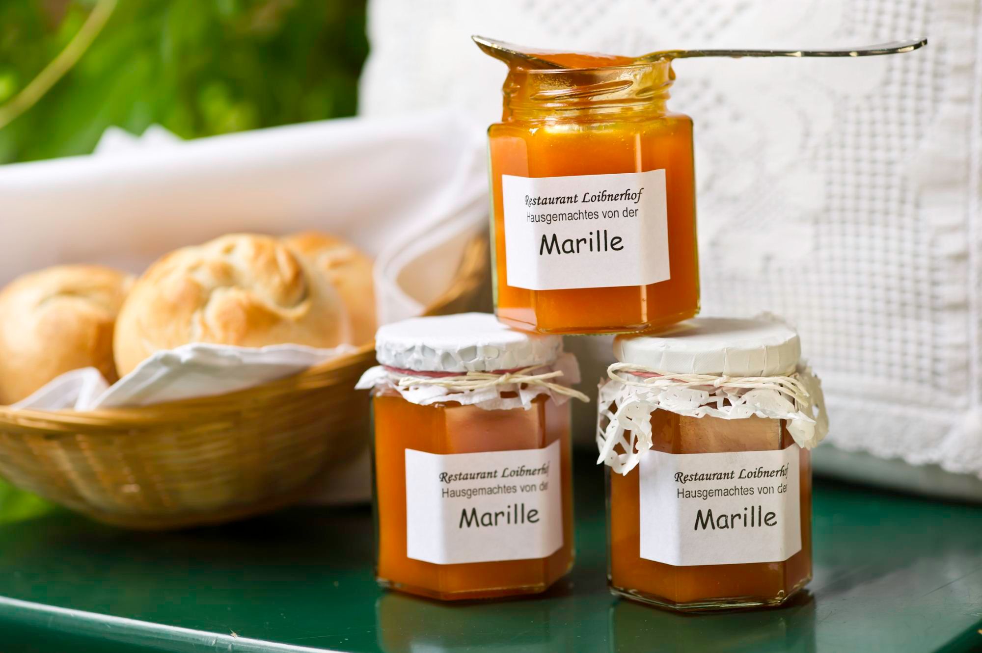Drei Gläser Marillenmarmelade vom Restaurant Loibnerhof neben einem Korb mit Brötchen.