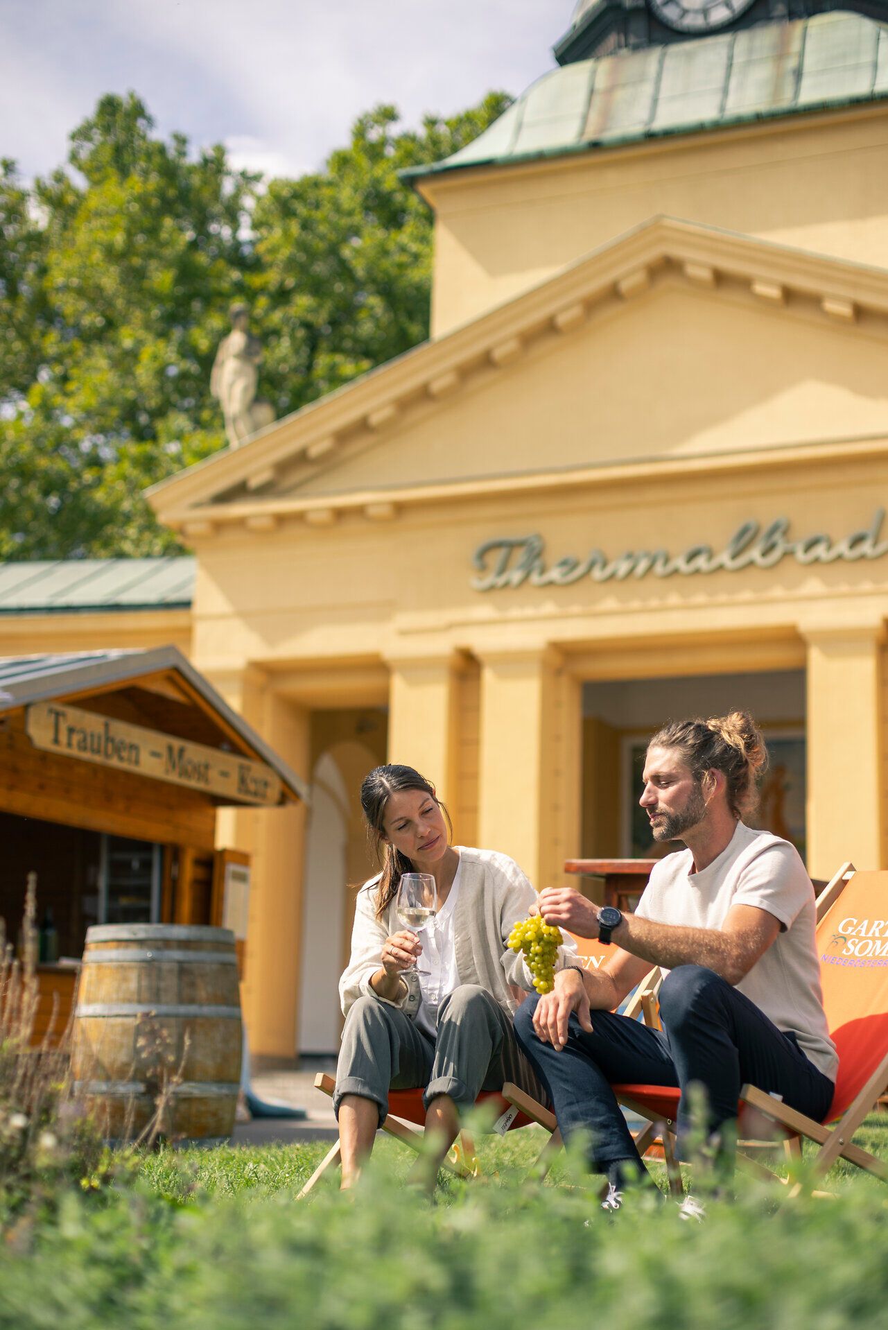 Vor dem Thermalbad Bad Vöslau sitzen ein Mann und eine Frau auf einem Liegestuhl vor dem Trauben-Most-Kur Stand. Der Mann hält grüne Trauben und die Dame ein Glas Wein in der Hand. 