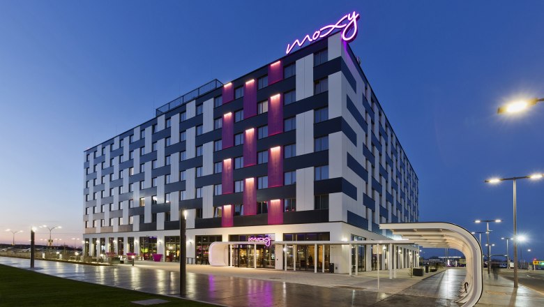 Außenansicht des Moxy Vienna Airport Hotels bei Dämmerung mit beleuchtetem Schriftzug.
