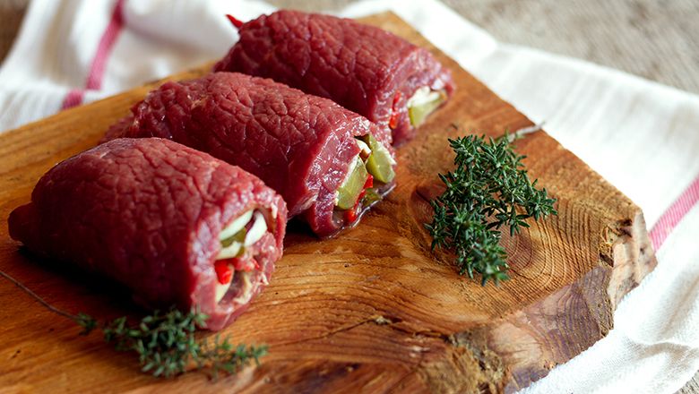 Drei gefüllte rohe Rinderrouladen auf einem Holzbrett mit Thymianzweigen.